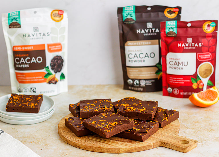 A wooden cutting board topped with squares of chocolate orange fudge surrounded by a stack of small white plates topped with fudge and bags of Navitas Organics Semi-sweet Cacao Wafers, Cacao Powder and Camu Powder, as well as a single orange slice.