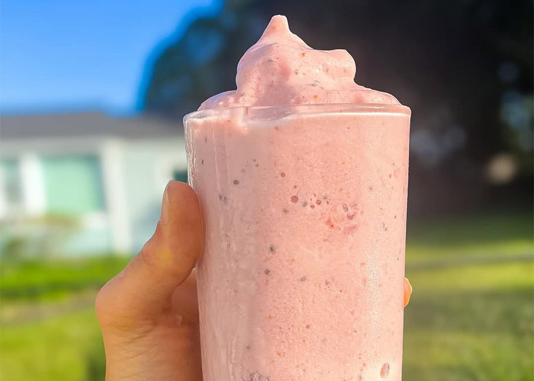 A person holding up a tall glass filled with a pink smoothie in the outdoors.