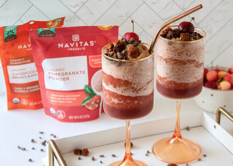 Two glasses filled with layered drinks on a white tray with bags of Navitas Organics Pomegranate Powder and Superfood Immunity Blend in the background surrounded by scattered cacao nibs and a bowl of cherries.