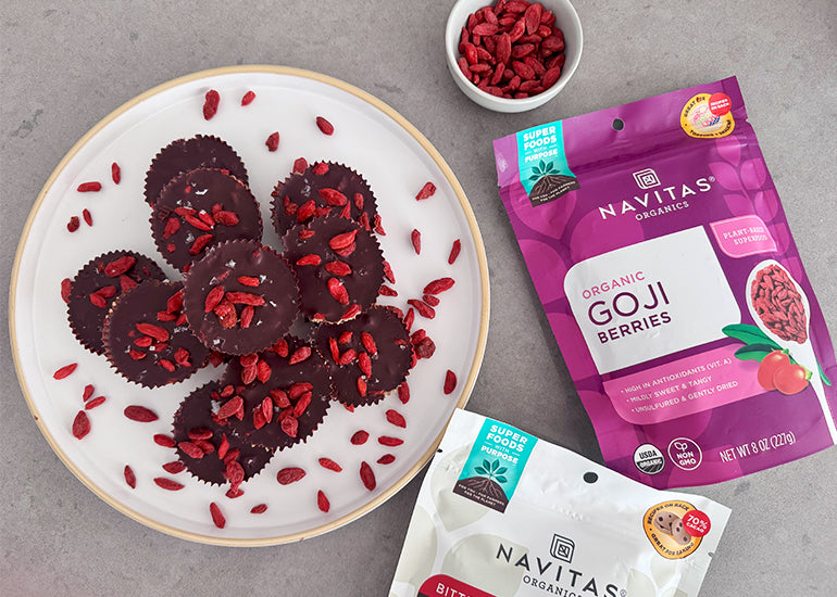 A plate of chocolate-covered peanut butter cups sprinkled with goji berries next to a small dish filled with goji berries, and bags of Navitas Organics Goji Berries and Bittersweet Cacao Wafers.