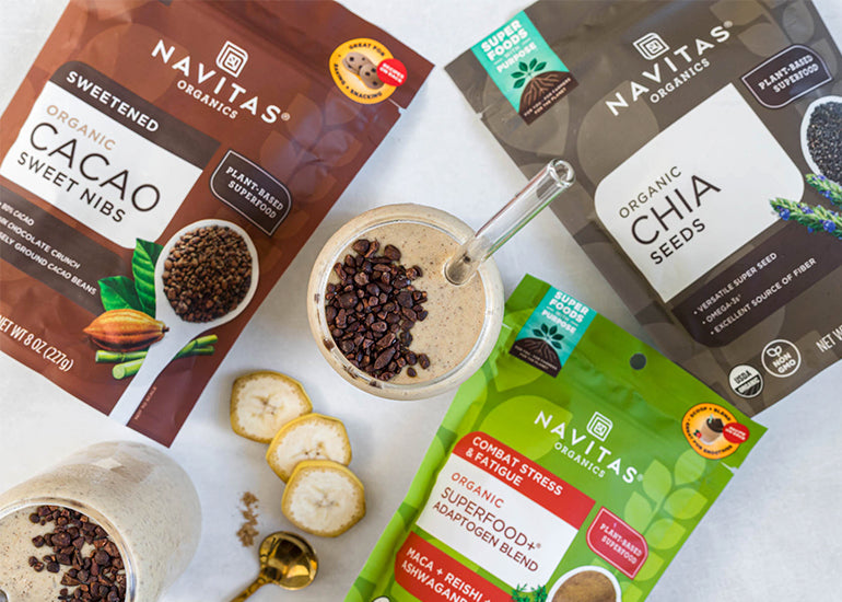 Overhead view of several Navitas Organics product packages including Cacao Sweet Nibs, Chia Seeds, and Superfood+ Adaptogen Blend, alongside a smoothie glass sprinkled with cacao nibs and slices of banana.