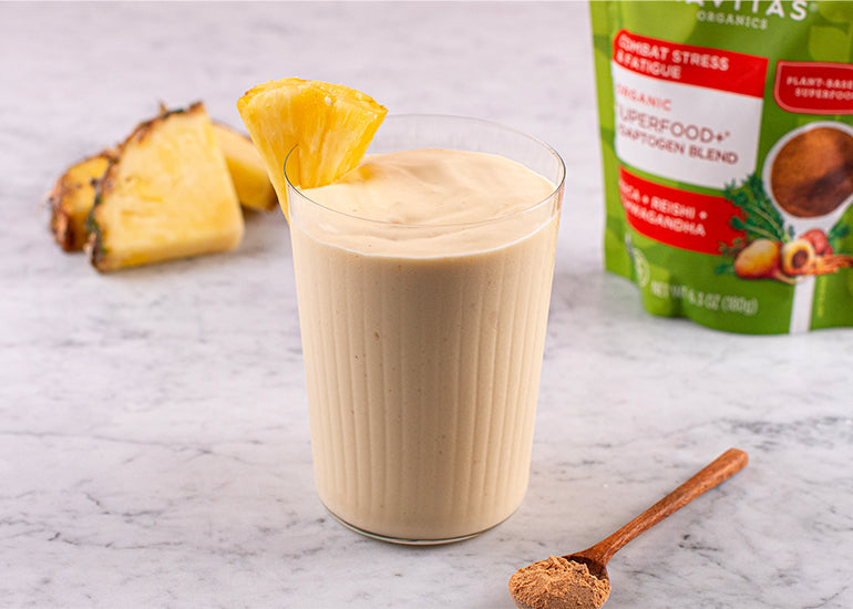 A glass of smoothie with a slice of pineapple on the rim, next to a cut pineapple piece, a spoonful of powder, and a bag of Navitas Organics Superfood+ Adaptogen Blend on a marble surface.