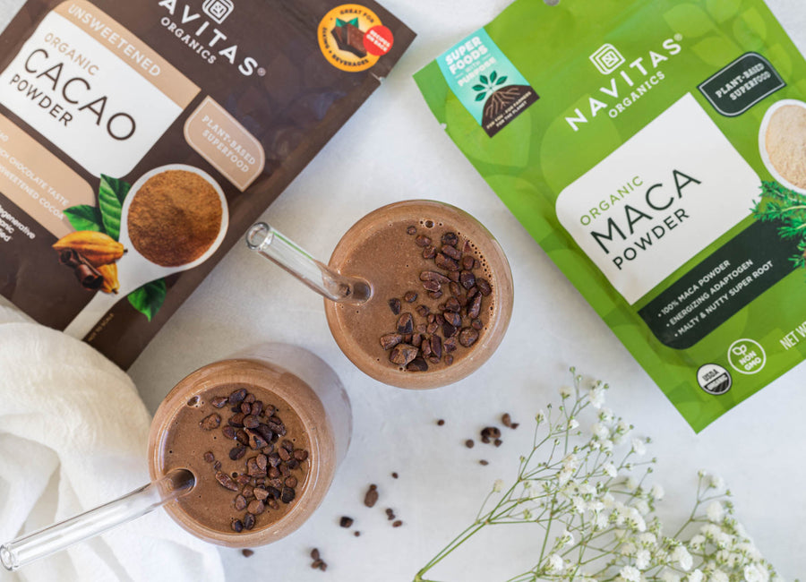 Two glasses of chocolate smoothie topped with cacao nibs, alongside bags of Navitas Organics Cacao Powder and Maca Powder.