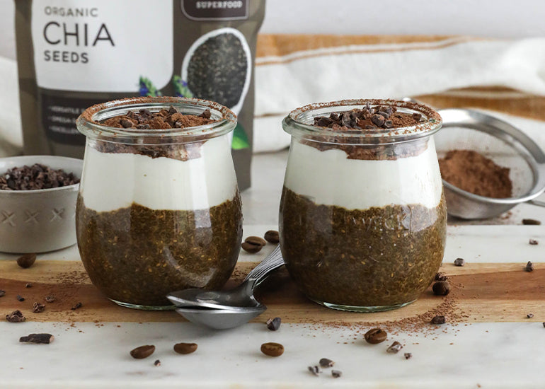 Two jars of layered chia pudding on a marble countertop, next to a bag of Navitas Organics Chia Seeds and scattered coffee beans around. Each jar is topped with white cream and Navitas Organics Cacao Nibs, accompanied by a spoon on the side.