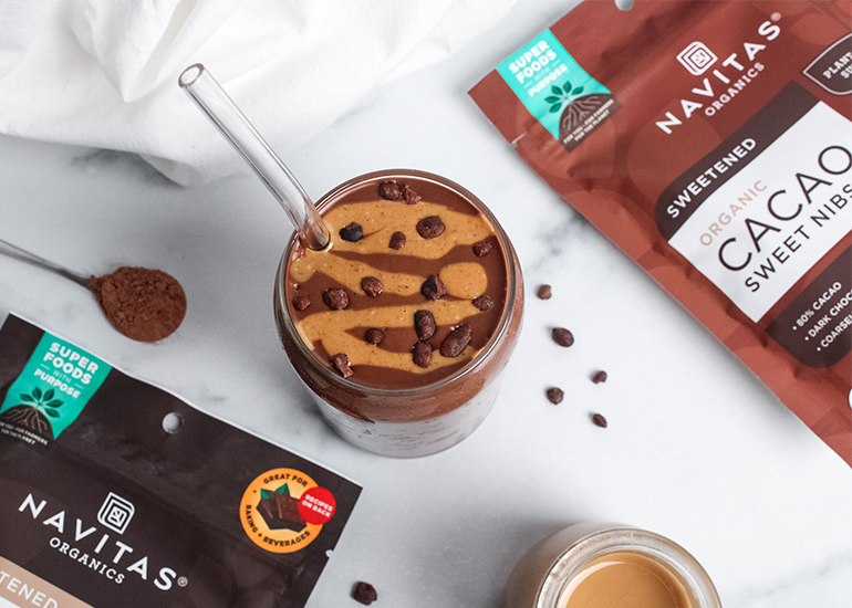 A chocolate peanut butter smoothie topped with cacao nibs, surrounded by Navitas Organics Cacao Powder and Cacao Sweet Nibs packaging on a marble surface.