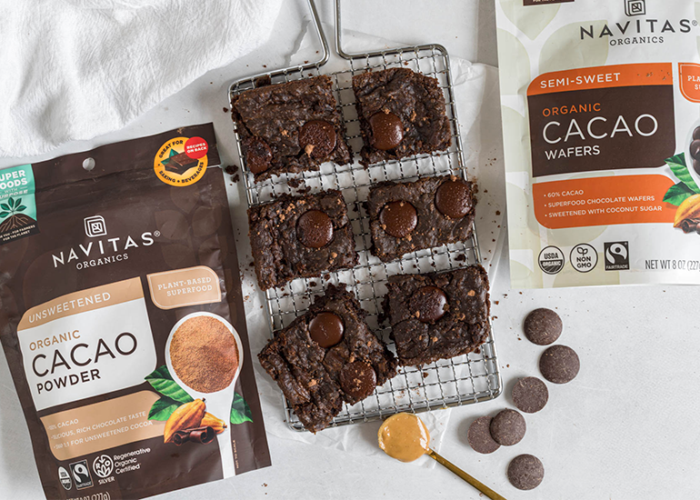 Freshly baked brownies with cacao wafers on a cooling rack beside bags of Navitas Organics Cacao Powder and Semi-sweet Cacao Wafers.