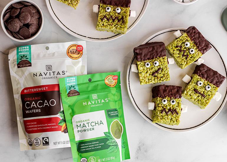 Plate of square, green matcha-flavored treats decorated to look like monsters with eyes and teeth, next to packages of Navitas Organics Bittersweet Cacao Wafers and Matcha Powder.