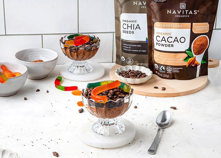 Chocolate chia pudding topped with cacao sweet nibs and gummy worm candies in a glass bowl, alongside a spoon and bags of Navitas Organics Chia Seeds and Cacao Powder.