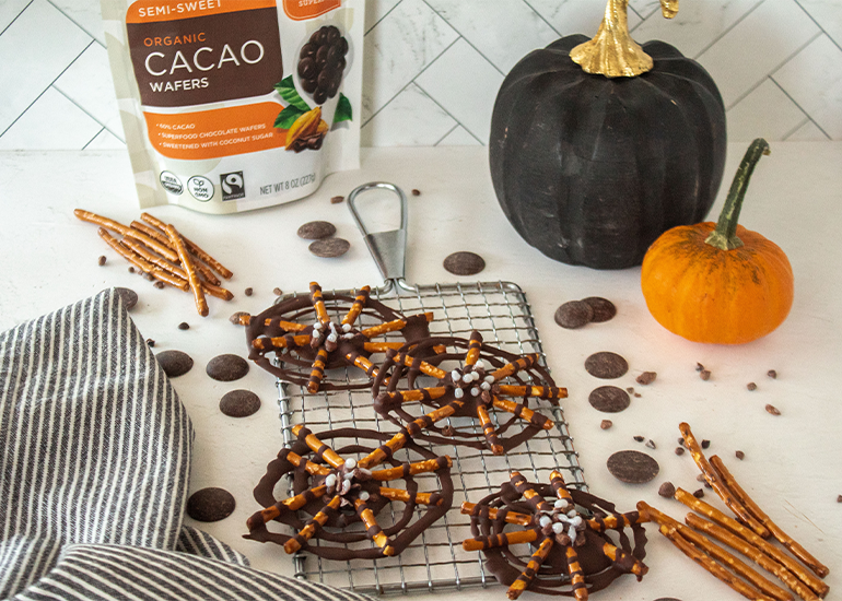 A kitchen counter setup featuring Navitas Organics Semi-sweet Cacao Wafers packaging, a black pumpkin, an orange pumpkin, and a cooling rack with chocolate-covered pretzels sprinkled with cacao nibs and dotted with white icing.