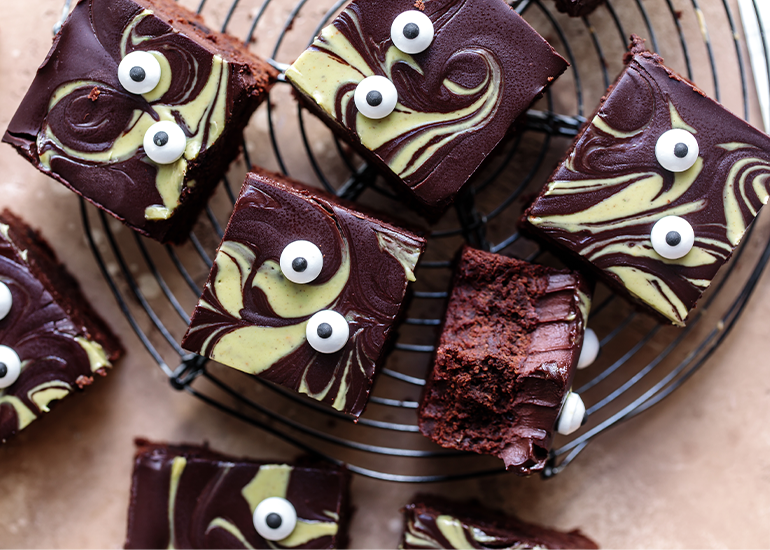 Chocolate brownies made with Navitas Organics Cacao Powder topped with a swirled pistachio and dark chocolate ganache made with pistachio butter and Navitas Organics Bittersweet Cacao Wafers, decorated with white candy eyes on a cooling rack.