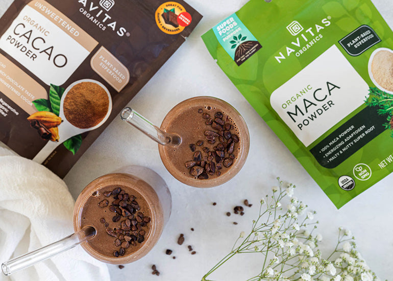 Two glasses of chocolate smoothie topped with cacao nibs, near packages of Navitas Organics Cacao Powder and Maca Powder, on a white surface with a subtle floral decoration.