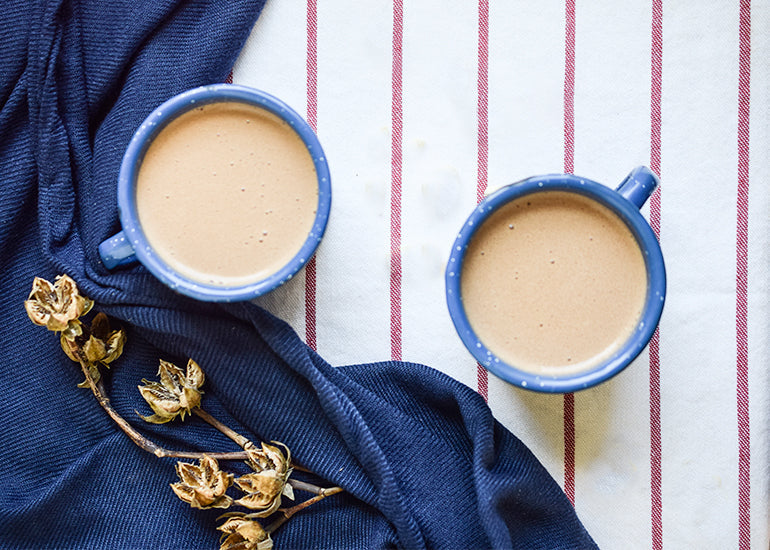 Two cups of coffee on a striped tablecloth next to a blue cloth and dried flowers.