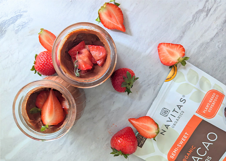 An overhead shot of two glass jars filled with chocolate mousse topped with strawberries surrounded by halved strawberries and a bag of Navitas Organics Semi-sweet Cacao Wafers.