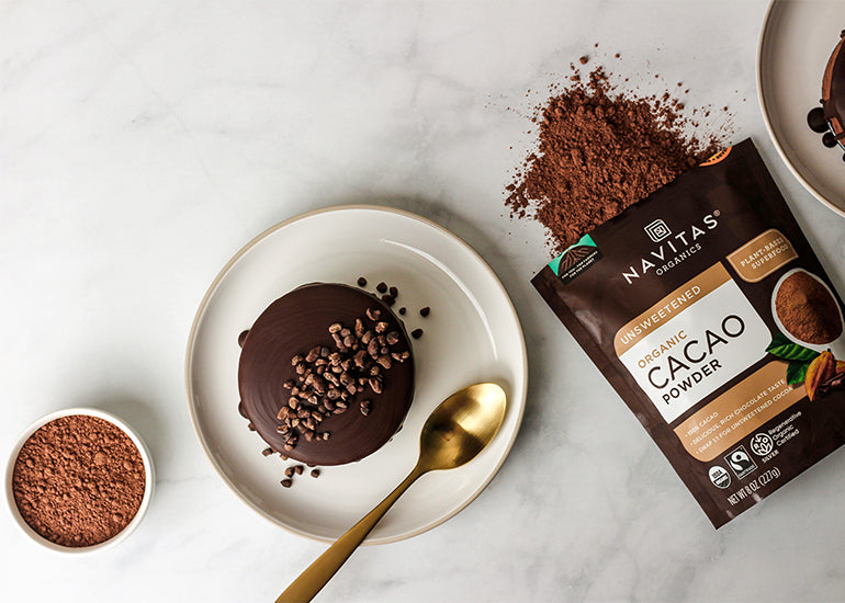 A mini chocolate mousse cake topped with Navitas Organics Cacao Nibs on a plate next to a dish and bag filled with Navitas Organics Cacao Powder