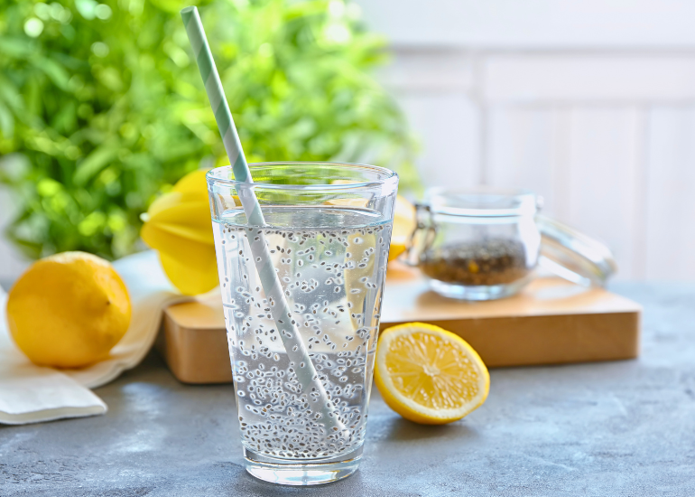 Glass of water with a straw, surrounded by sliced lemons and a jar of chia seeds on a kitchen counter.
