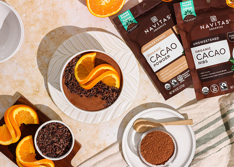 A chocolate orange smoothie bowl surrounded by Navitas Organics Cacao Powder and Cacao Nibs in dishes and in bags