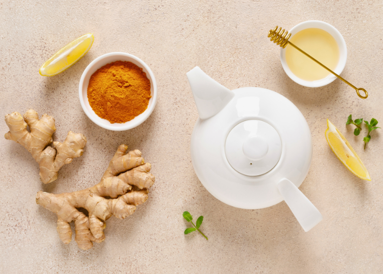 A tea kettle, dish filled with turmeric powder, turmeric root, slices of lemon, and a mug topped with a honey stirrer
