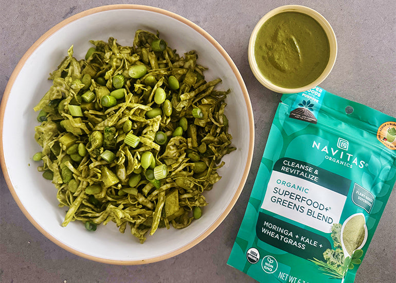 A bowl of tossed cabbage, cucumbers and edamame next to a bag of Navitas Organics Superfood+ Greens Blend and a small dish filled with homemade green goddess dressing