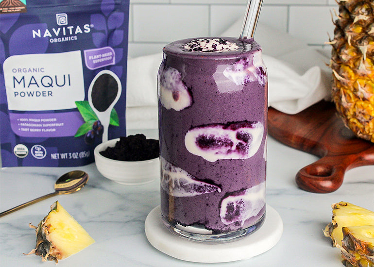 A tall glass filled with a maqui smoothie surrounded by pineapple and a bag of Navitas Organics Maqui Powder.