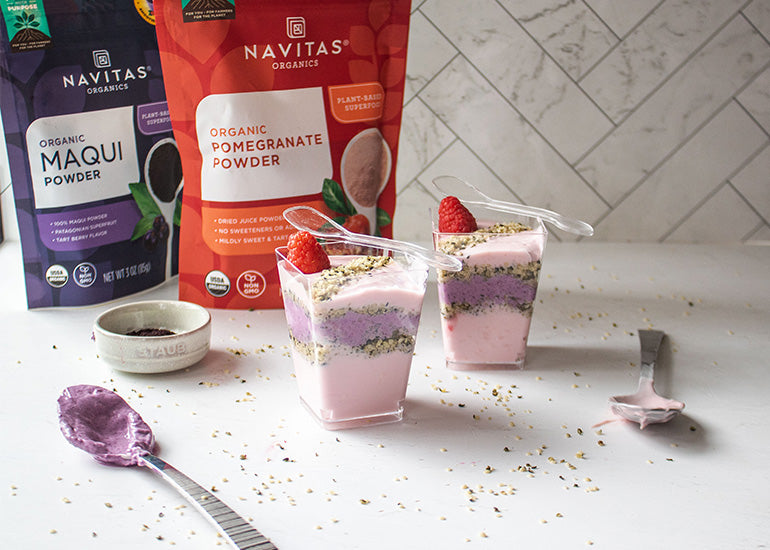 Two pink and purple layered parfaits topped with hemp seeds in front of bags of Navitas Organics Maqui Powder and Pomegranate Powder, displayed on a kitchen counter with decorative spoons and raspberries.