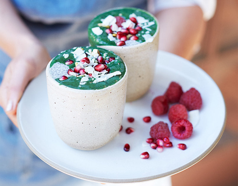 A woman holding a tray with two green smoothies made with Navitas Organics Wheatgrass Powder, topped with fresh raspberries and shaved coconut