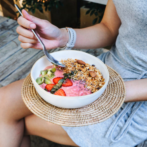 A woman in a gray dress sitting on a wooden deck holding a spoon above a berry, granola and seed-topped smoothie bowl.