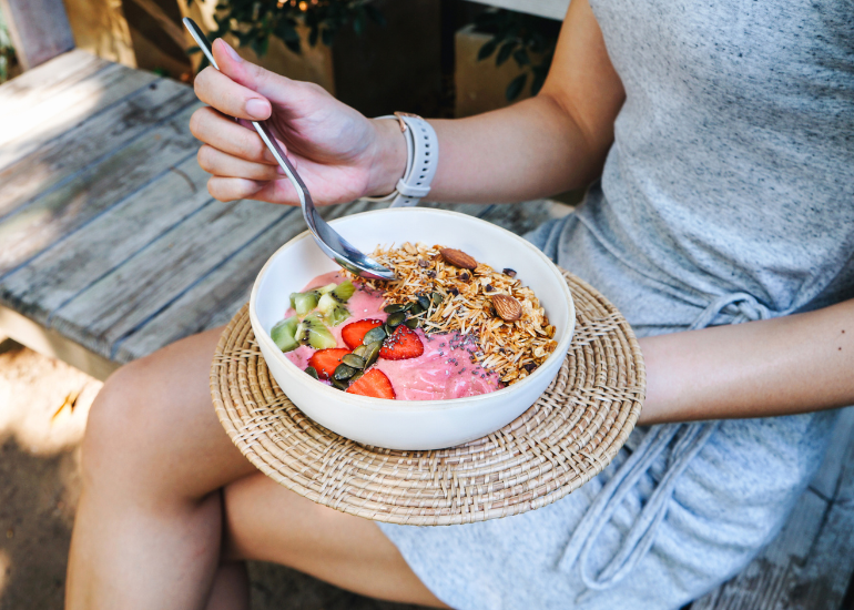A woman in a gray dress sitting on a wooden deck holding a spoon above a berry, granola and seed-topped smoothie bowl.