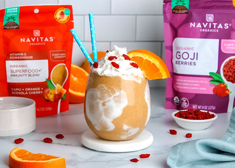 An orange smoothie swirled with coconut yogurt topped with whipped cream, goji berries and an orange wedge surrounded by a small dish with goji berries and bags of Navitas Organics Superfood+ Immunity Blend and Goji Berries.
