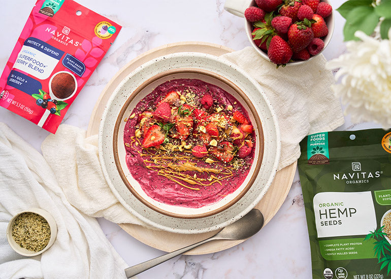 An overhead shot of a smoothie bowl topped with berries, nut butter and hemp seeds surrounded by a dish with fresh strawberries, a dish filled with hemp seeds and bags of Navitas Organics Hemp Seeds and Superfood+ Berry Blend.