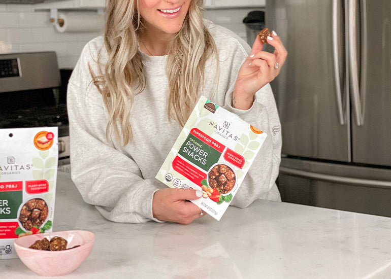 A person holding a bag of Navitas Organics Superfood PB&J Power Snacks, smiling while sitting at a kitchen counter with a bowl of snacks and another product bag visible.