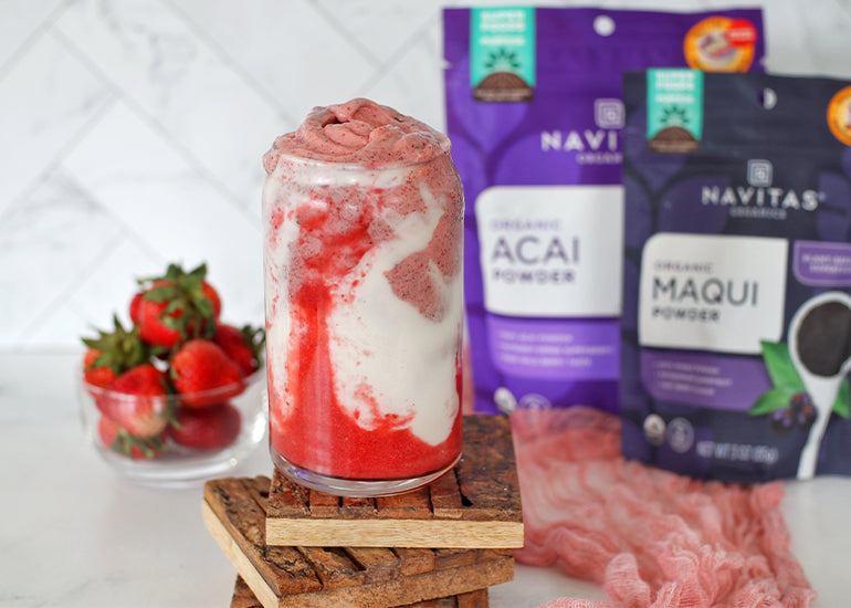 A pink swirled smoothie in a tall glass on a kitchen counter in front of a small dish filled with fresh strawberries and bags of Navitas Organics Maqui Powder and Acai Powder.