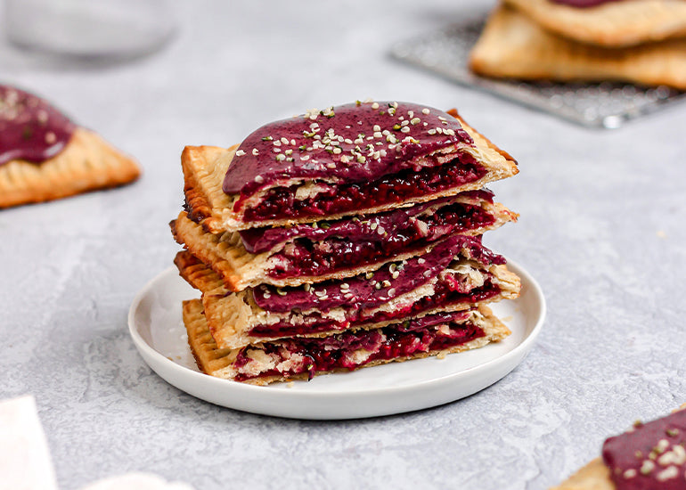 A stack of homemade berry pop tarts on a plate topped with hemp seeds surrounded by additional individual pop tarts