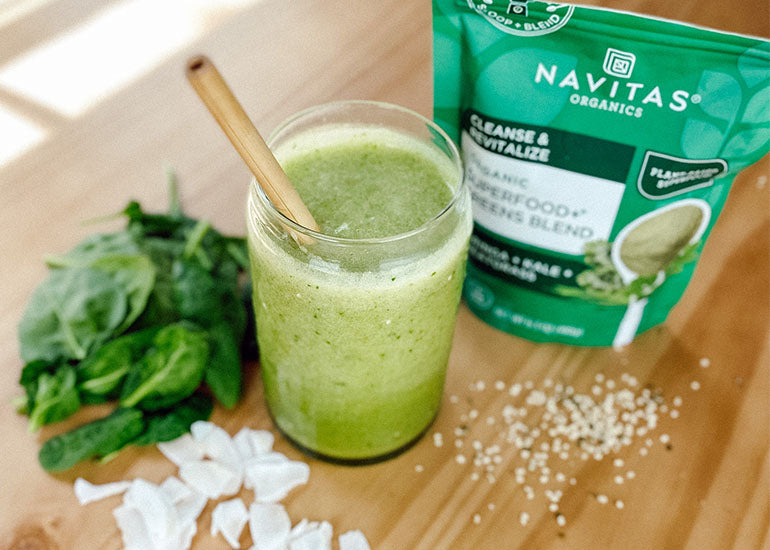 A green smoothie made with Navitas Organics Superfood+ Greens Blend on a table with hemp seeds, fresh spinach and shaved coconut.