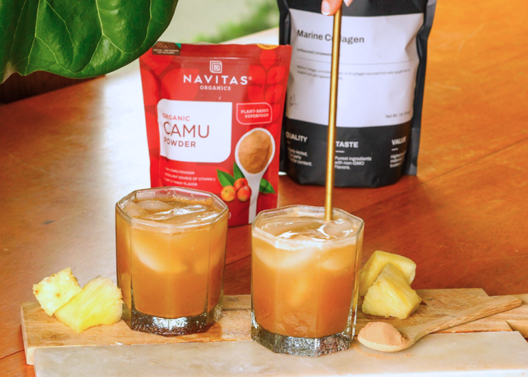 Two glasses of a beverage with ice, garnished with pineapple slices on a wooden board, next to packages of Navitas Organics Camu Powder and True Nutrition Marine Collagen.