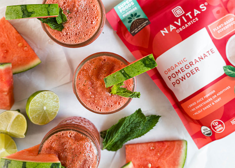 Two glasses of watermelon slushie garnished with mint leaves, surrounded by slices of watermelon, a lime, and a bag of Navitas Organics Pomegranate Powder.