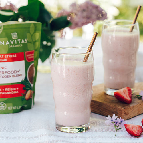 Two pink smoothies in tall glasses on a clothed table next to sliced strawberries, flowers and a bag of Navitas Organics Superfood+ Adaptogen Blend.