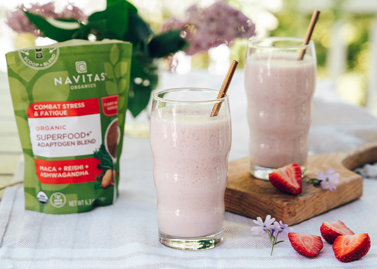 Two pink smoothies in tall glasses on a clothed table next to sliced strawberries, flowers and a bag of Navitas Organics Superfood+ Adaptogen Blend.