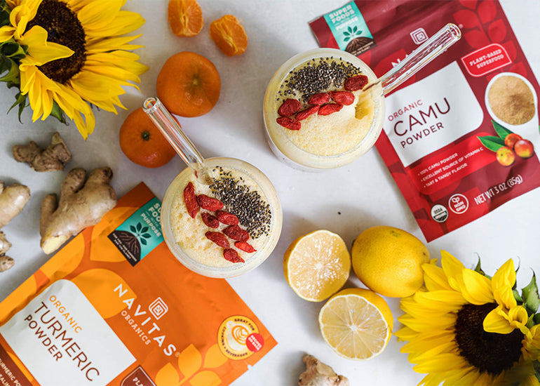 An overhead shot of two tall citrus smoothies topped with goji berries and chia seeds surrounded by sunflowers, lemons, ginger, a bag of Navitas Organics Camu Powder, and a bag of Navitas Organics Turmeric Powder.
