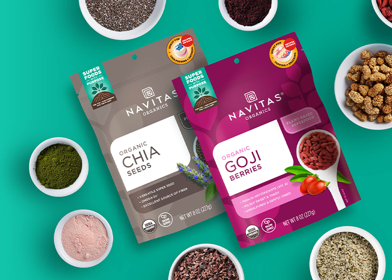 A bag of Navitas Organics Chia Seeds and Goji Berries surrounded by small dishes filled with various superfood berries, seeds and powders, all against a teal background.
