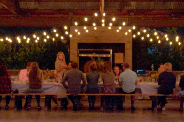 Long table with strings of lights overhead with people eating together.