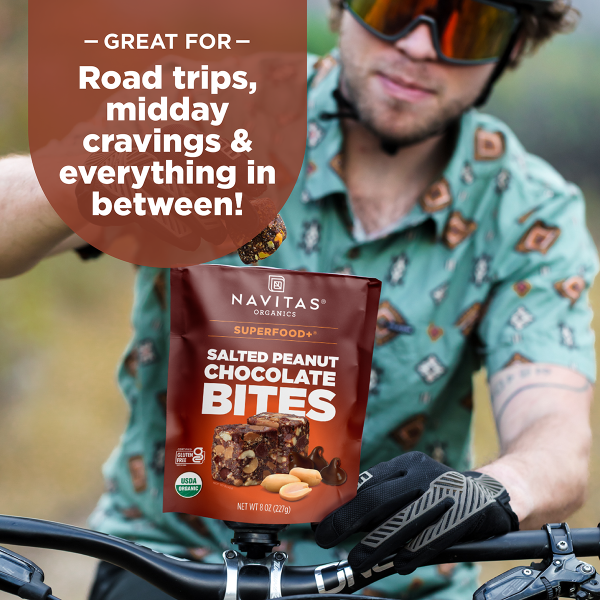 Person cycling on a trail while holding a package of Navitas Organics Superfood Salted Peanut Chocolate Bites, with text promoting it as great for road trips, cravings, and everyday moments.