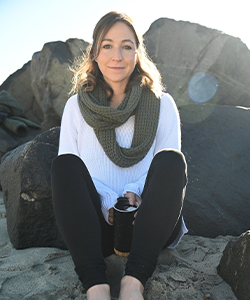 Certified Nutritionist and Ayurvedic Wellness Counselor, Emily Ziedman, MS, CN sitting on rocks at a beach holding a thermos with a scarf wrapped around her neck.