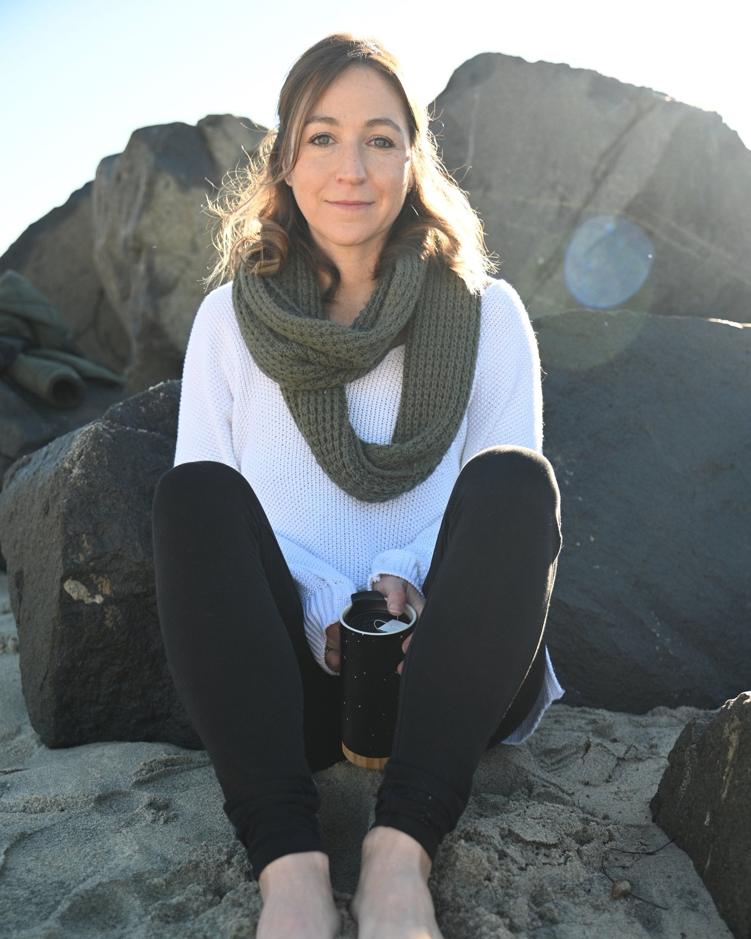 Certified Nutritionist and Ayurvedic Wellness Counselor, Emily Ziedman, MS, CN sitting on rocks at a beach holding a thermos with a scarf wrapped around her neck.