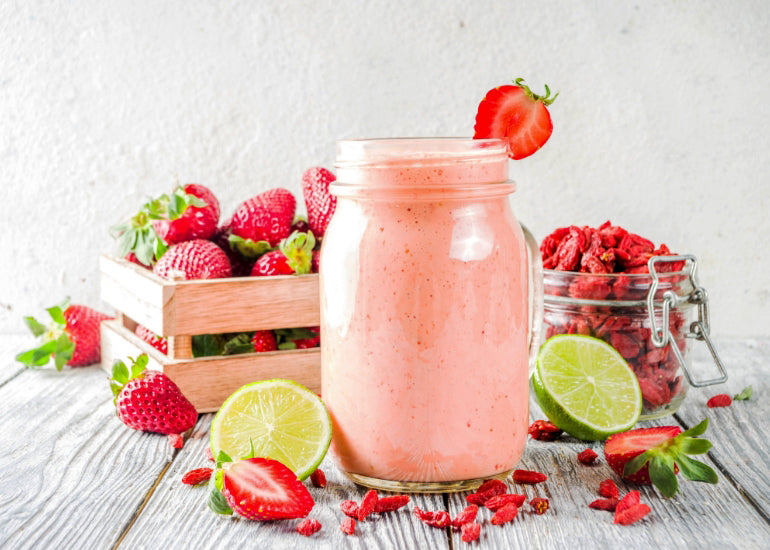 A smoothie in a glass jar surrounded by dried goji berries, limes and fresh strawberries