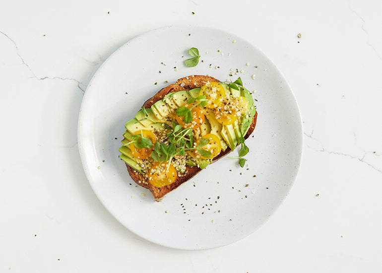 Avocado toast on a plate, topped with fresh herbs, tomatoes, and Navitas Organics Hemp Seeds and Chia Seeds