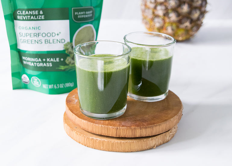 Two green drinks made with Navitas Organics Superfood+ Greens Blend in glasses on wooden boards 