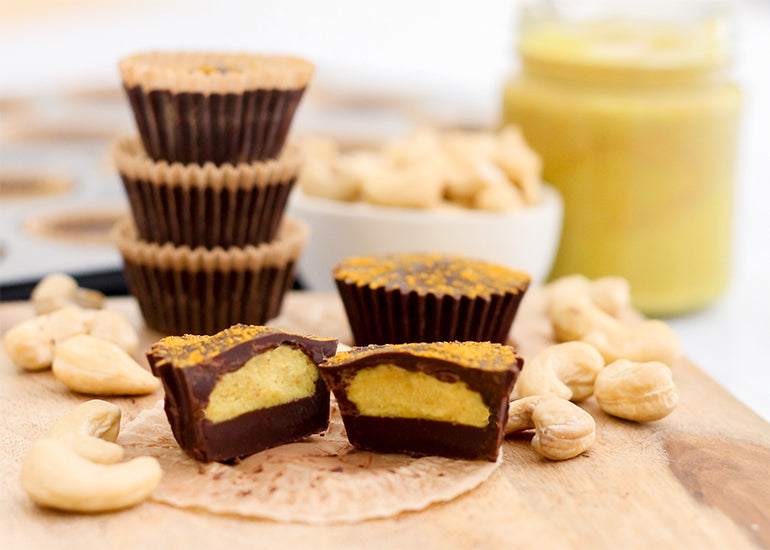Stacks of chocolate golden milk cashew butter cups made with Navitas Organics Cacao Butter, Cacao Powder, Cashew Nuts and Turmeric Powder on a wooden cutting board surrounded by cashews 