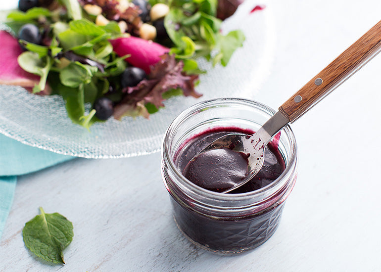 A glass jar filled with dark purple vinaigrette made with Navitas Organics Maqui Berry Powder, alongside a plate filled with green salad
