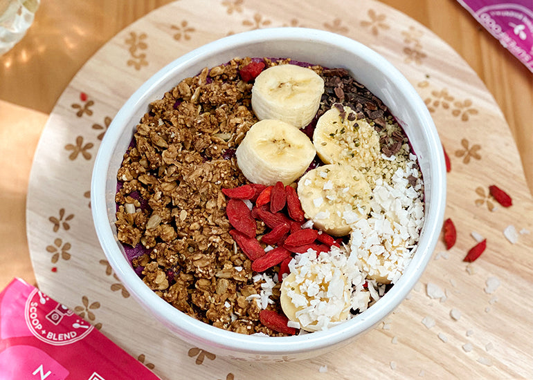 A smoothie bowl made with Navitas Organics Superfood+ Berry Blend, topped with banana slices, coconut flakes, granola, and Navitas Organics Goji Berries, Hemp Seeds and Chia Seeds