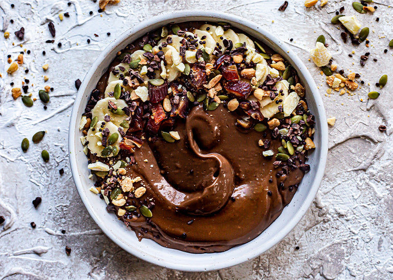 A chocolate smoothie bowl made with Navitas Organics Cacao Powder, Cacao Nibs and Hemp Seeds, topped with banana slices, nuts and seeds